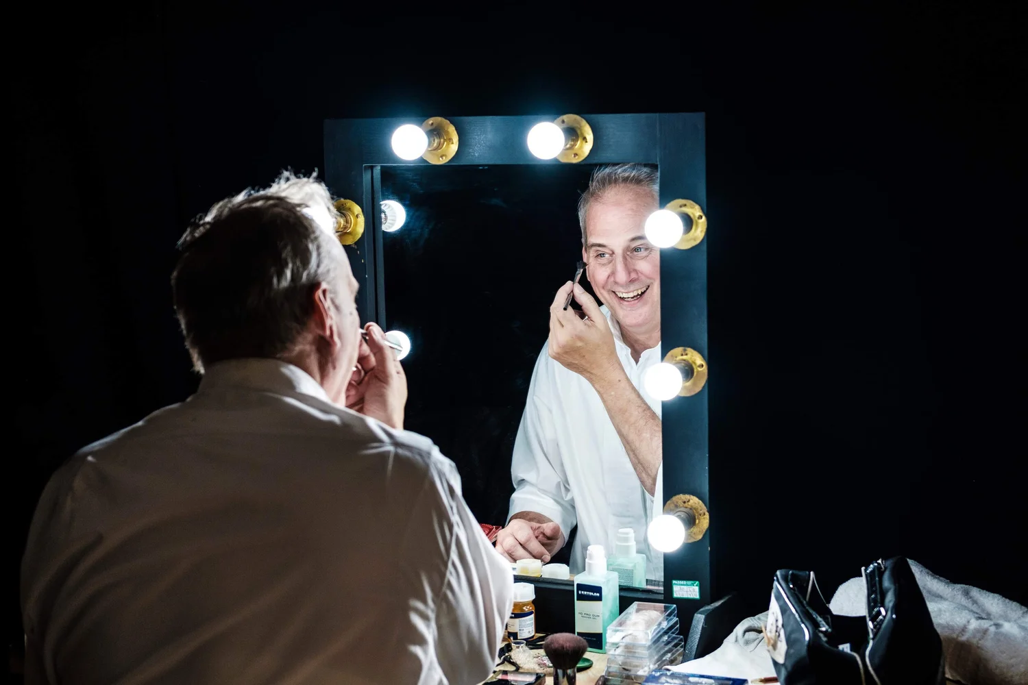 David Williamson smiling into a lighted dressing-room mirror while getting ready for a show